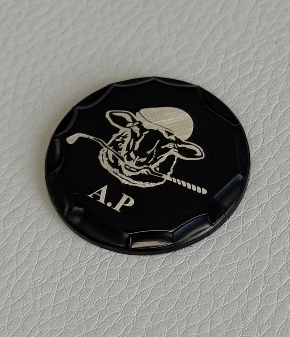 Black custom ball marker engraved with a cow golfer logo and initials A.P