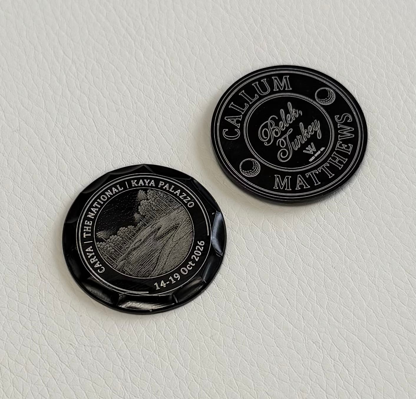 Two custom engraved black golf ball markers personalised with a name, golf course design and date